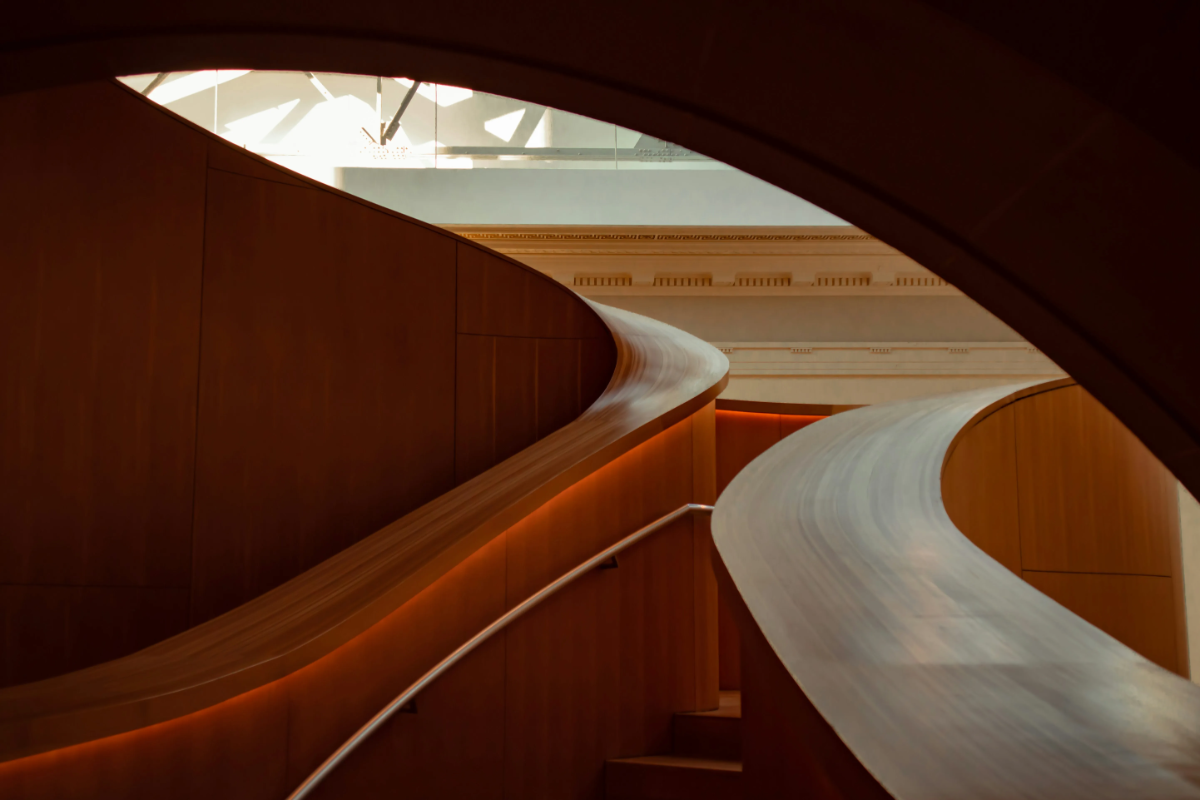 Curving, sculptural wooden staircase and ramp inside a modern museum, accented by warm indirect lighting.