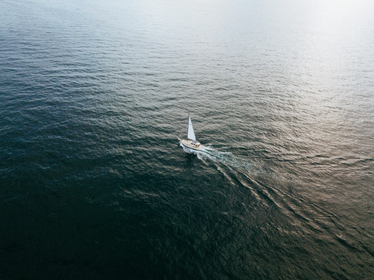 Aerial view of lone white-sail yacht carving gentle wake across open deep-blue ocean.
