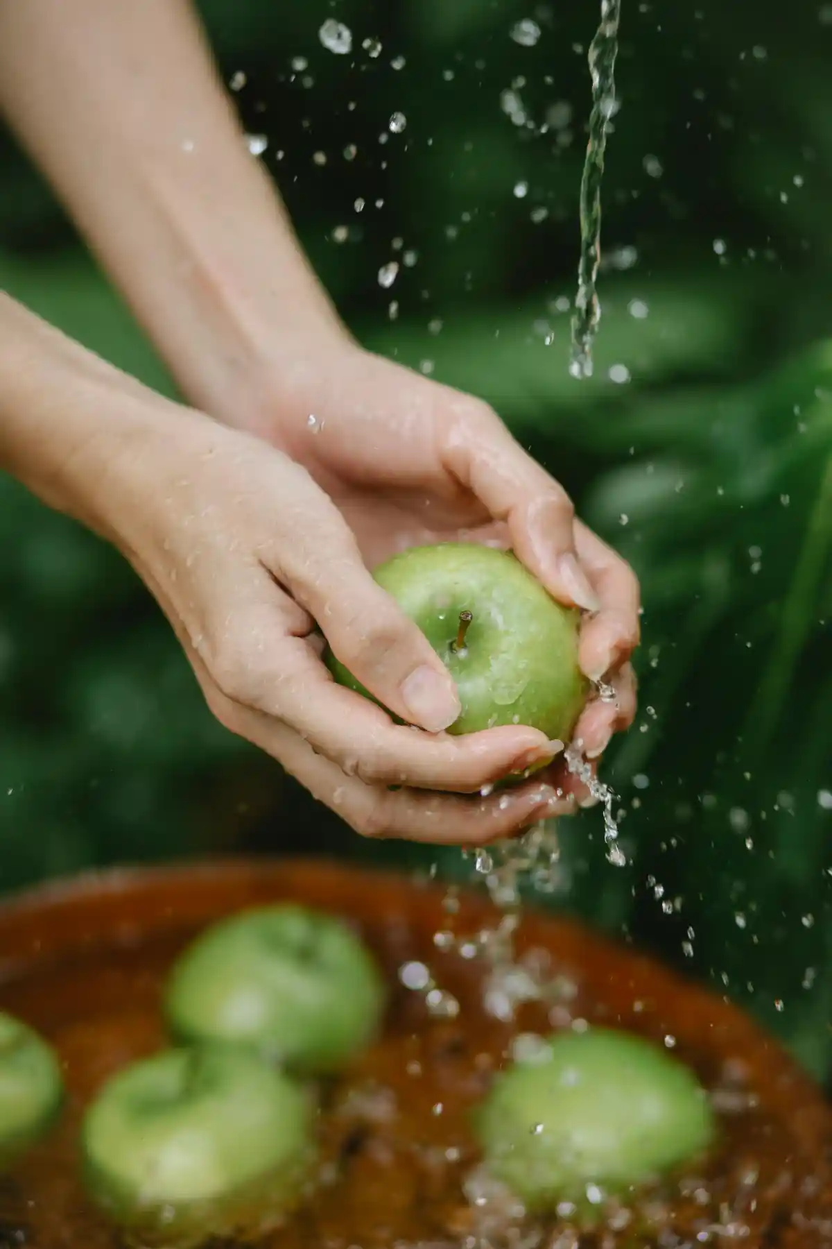 Two hands rinsing a green apple under splashing water with blurred garden background.
