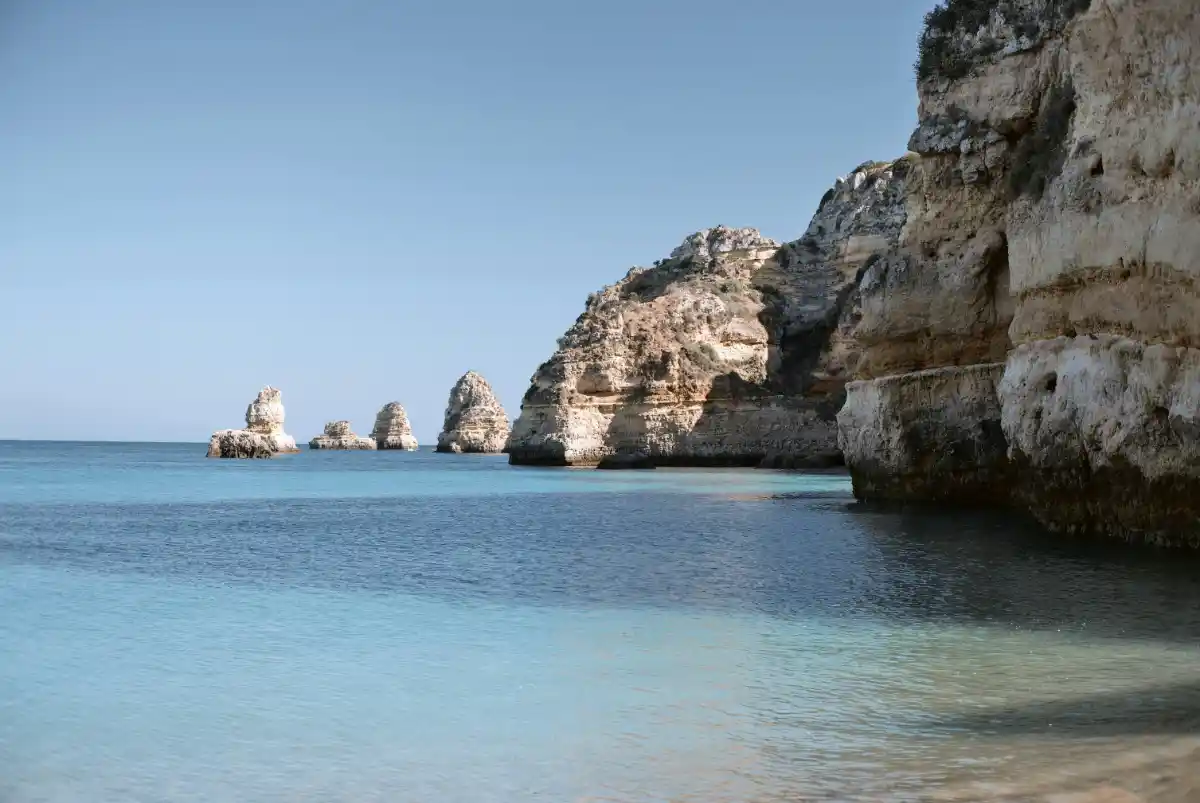 Limestone sea stacks rise from a calm, pale-blue bay on the coast of Algarve.