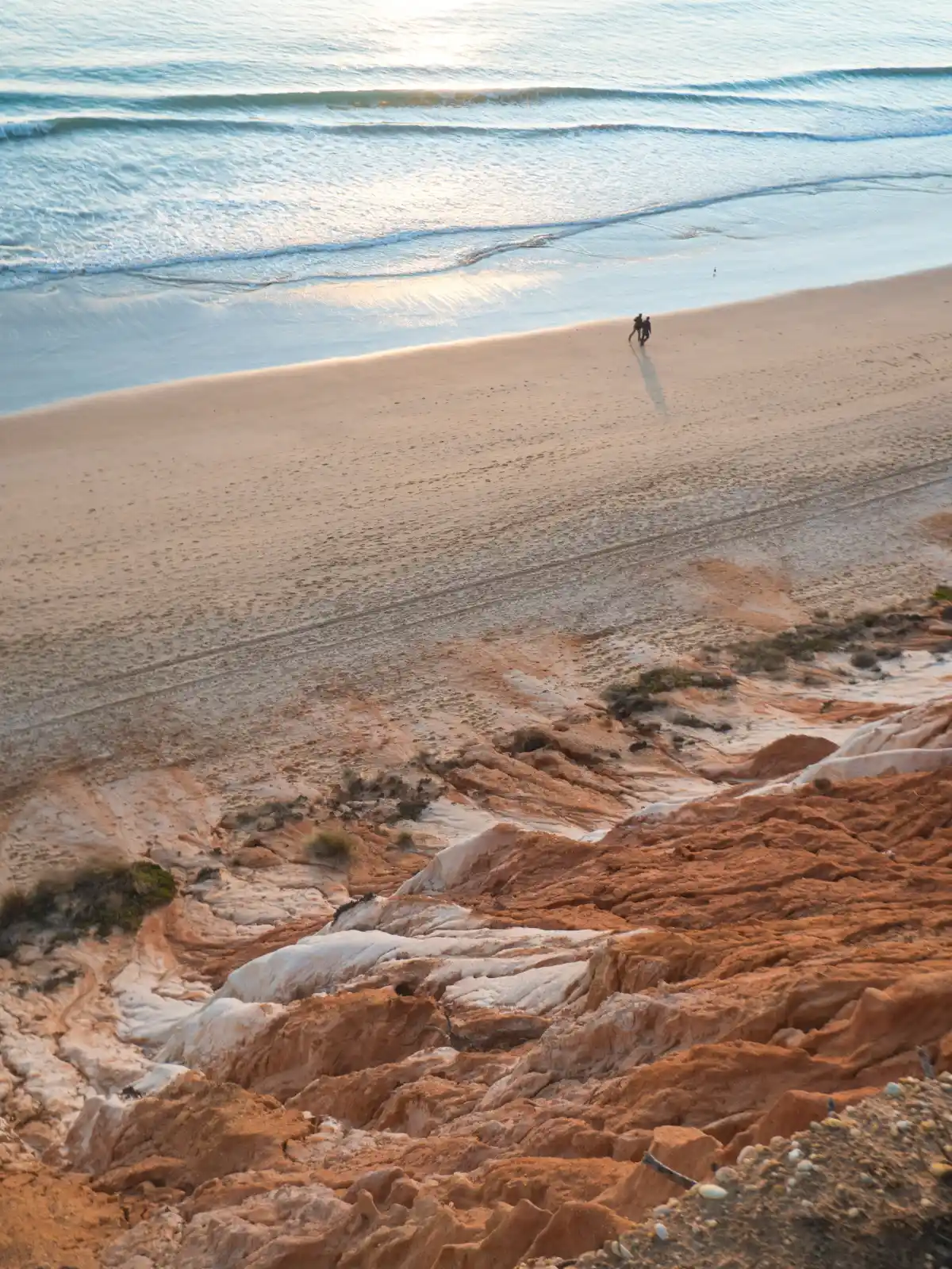 Couple strolls a quiet sunset shoreline beneath striking red-and-white sandstone cliffs of Praia da Falésia.