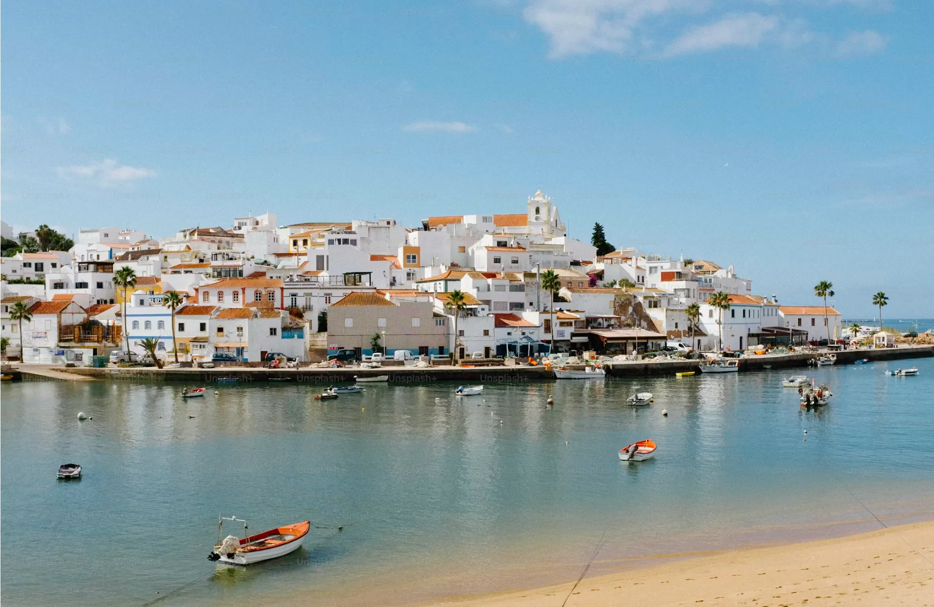 Whitewashed hillside town of Lagos reflected in calm harbor, dotted with small fishing boats and sandy shore.