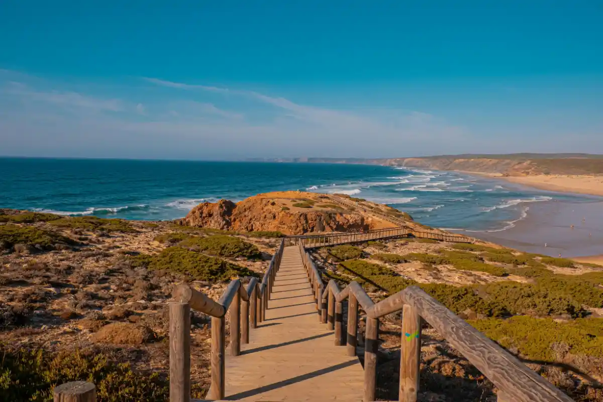 Long wooden boardwalk crosses rolling dunes toward the wide surf of Praia da Bordeira on Algarve’s wild west coast.