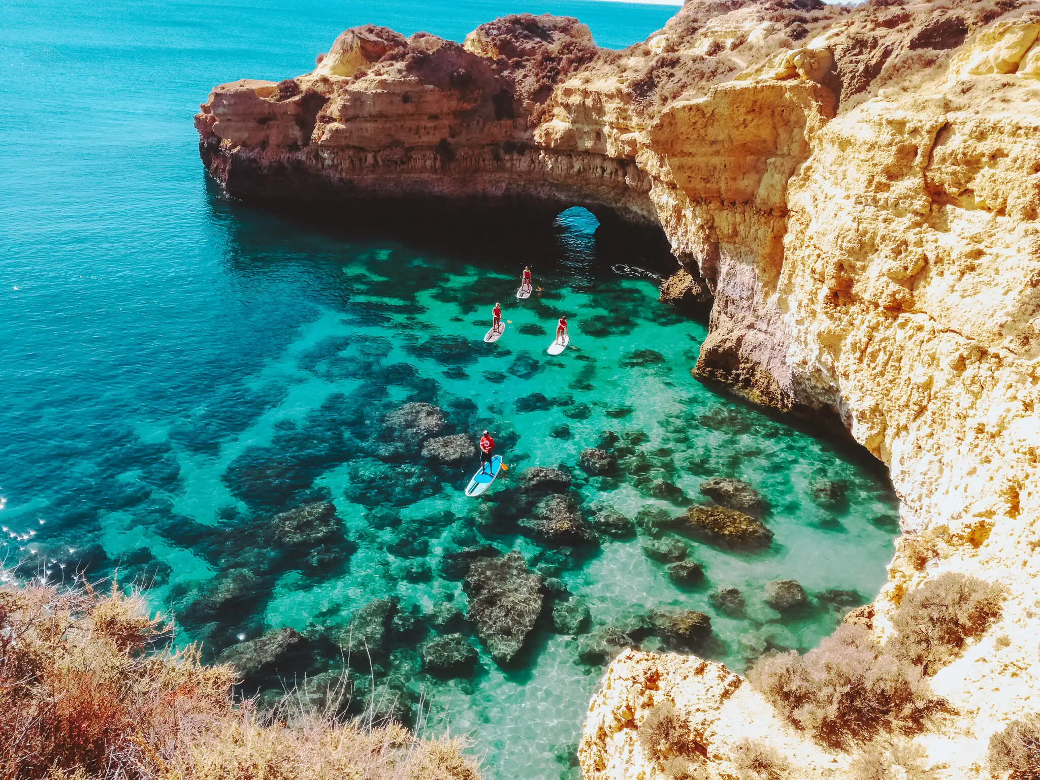 Stand-up paddleboarders glide over clear turquoise cove beneath limestone arch at Praia de São Rafael.
