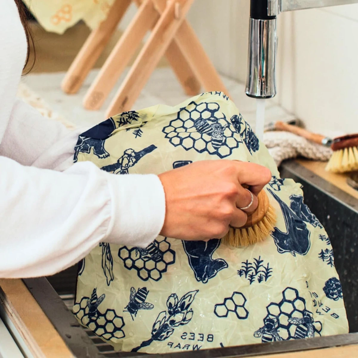 Close-up of hands scrubbing a patterned beeswax food wrap under a running tap with a natural bristle dish brush.