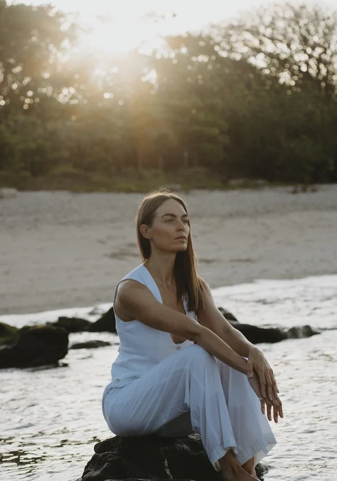 Woman seated on coastal rocks at sunset in white outfit, calm ocean setting.