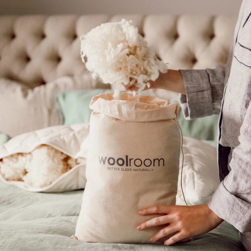 A hand pours raw wool fleece into a Woolroom bag set on sage green bed linens.