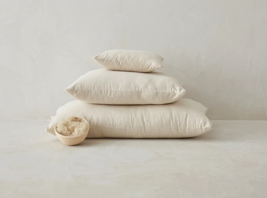 Three Futon Shop wool pillows in graduated sizes are stacked beside a small bowl of raw wool.