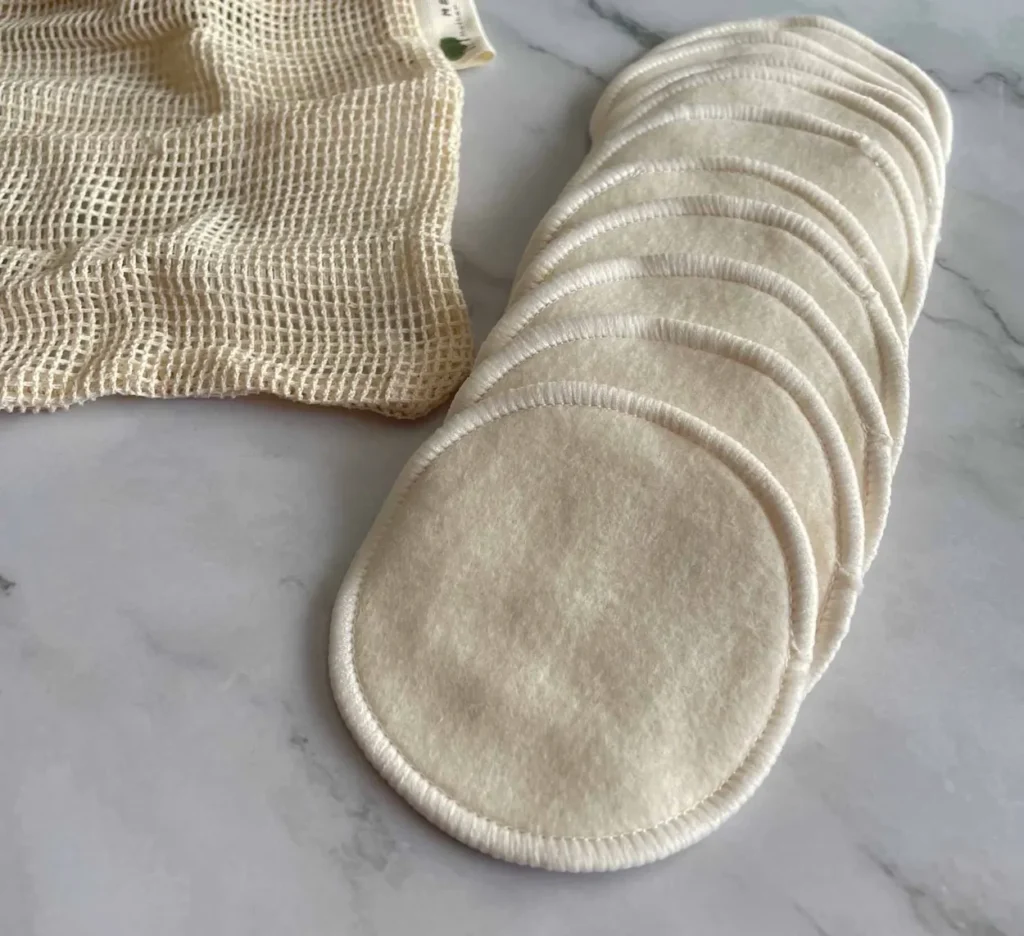 A fanned row of unbleached cotton and hemp facial rounds beside an open mesh laundry bag on marble.