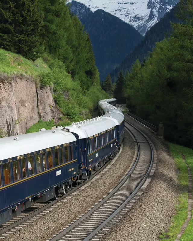 The navy-and-cream carriages curve through a forested alpine valley beneath snow-capped peaks.