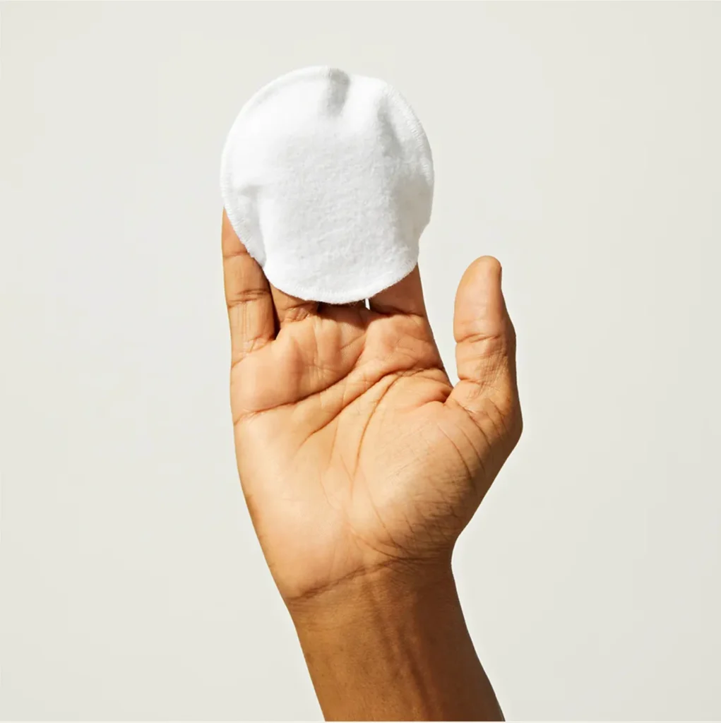 A single white reusable cotton round balanced on a raised open palm against a pale grey ground.