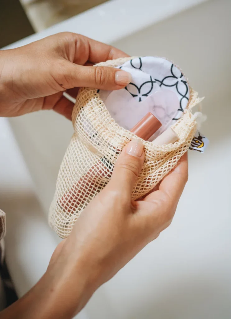 Hands open a cotton mesh bag to reveal Superbee patterned reusable rounds and a lip product inside.