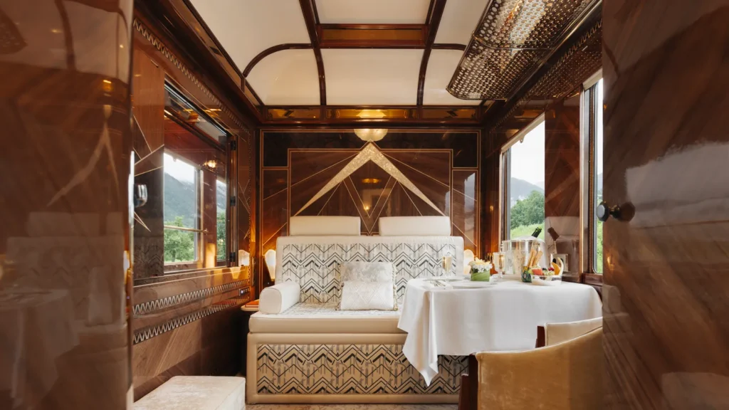 A Grand Suite cabin with Art Deco marquetry panels, a dressed dining table, champagne on ice, and mountain views beyond