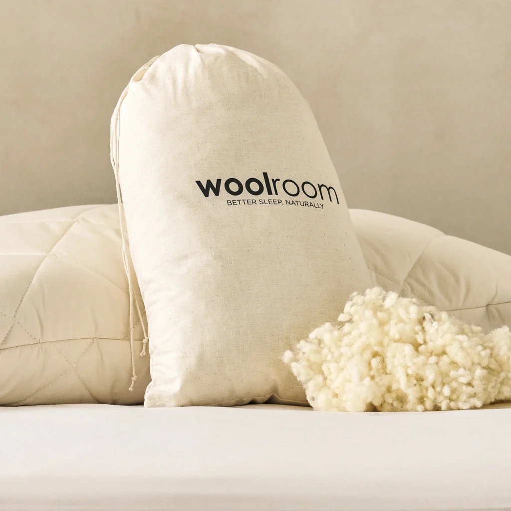 A Woolroom drawstring bag rests against quilted pillows beside a tuft of raw fleece.