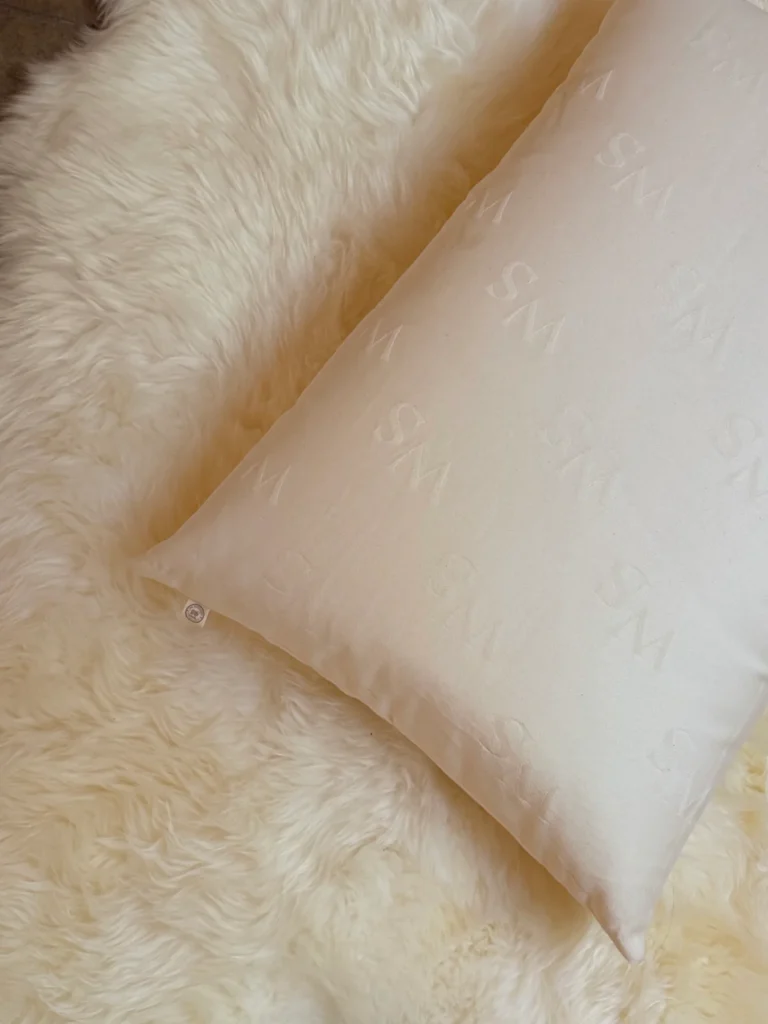 A Woolshire pillow embossed with a repeating monogram rests diagonally on a plush sheepskin rug.