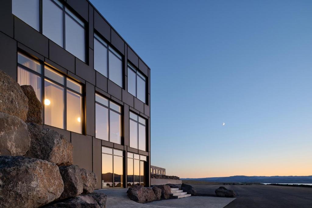 The dark-clad facade of Black Sand Hotel at dusk, its lit windows set among lava boulders beneath a crescent moon.