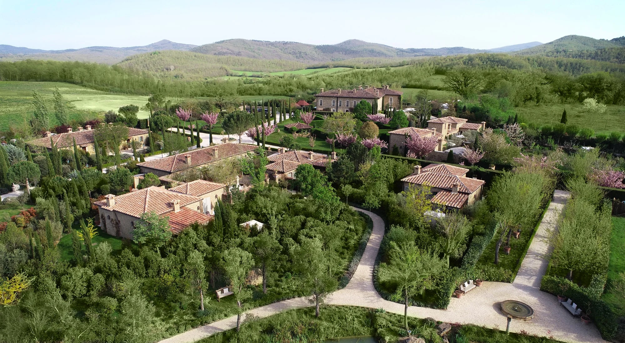 An aerial view of the Borgo Santo Pietro estate, with terracotta roofs, gravel paths, and gardens amid wooded hills.