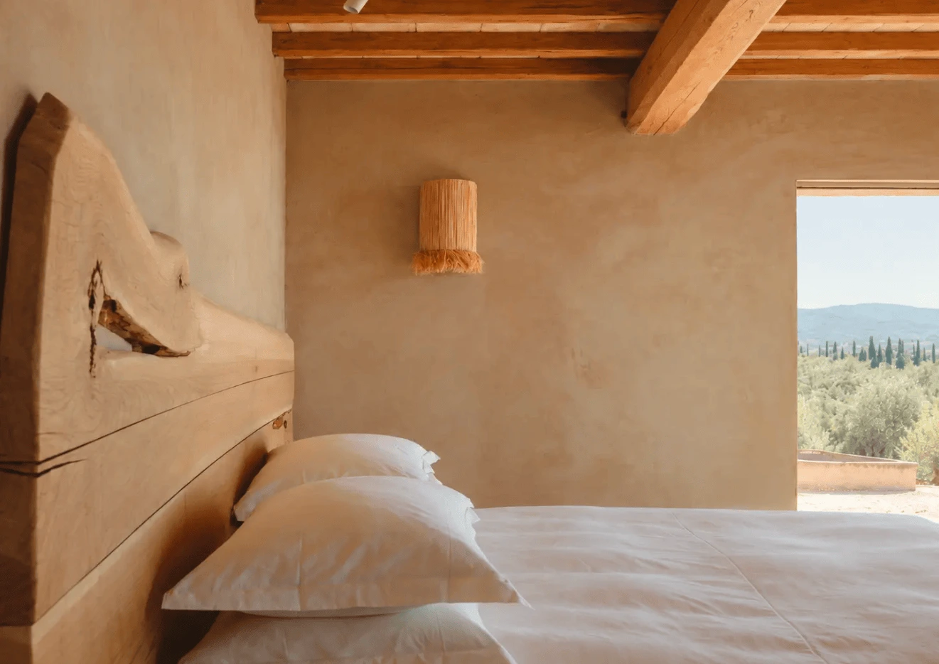 A live-edge timber headboard in a plaster bedroom at Conti di San Bonifacio, open to olive groves and cypresses.