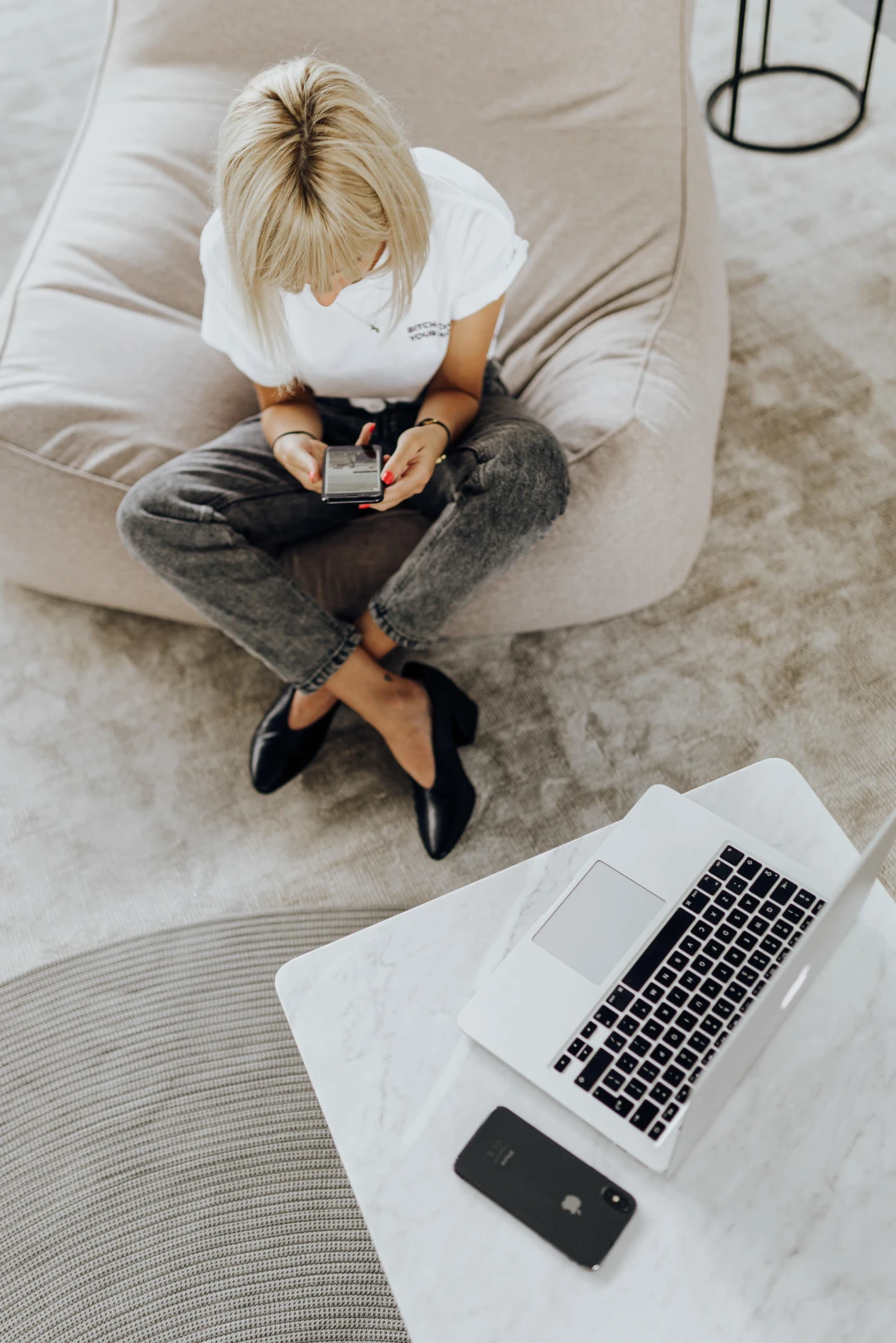 Woman sitting on a floor cushion checking her phone near a minimalist desk with a laptop.