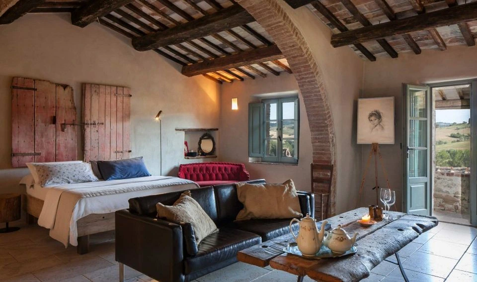 A beamed suite at Follonico with a brick arch, leather sofa, antique door headboard, and a tea service on a rustic table.