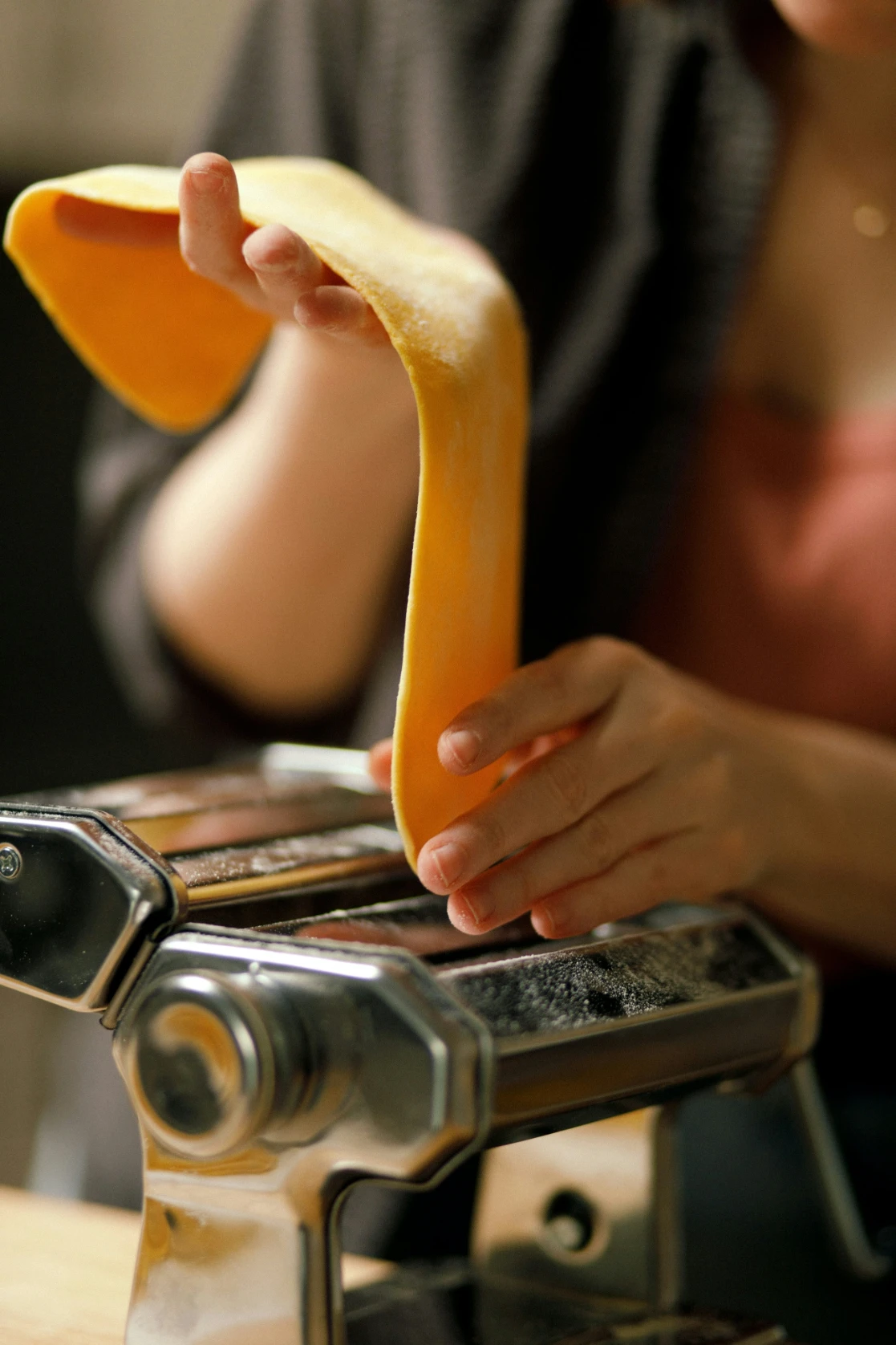 Hands lift a golden sheet of fresh egg pasta from a flour-dusted hand-crank pasta machine.