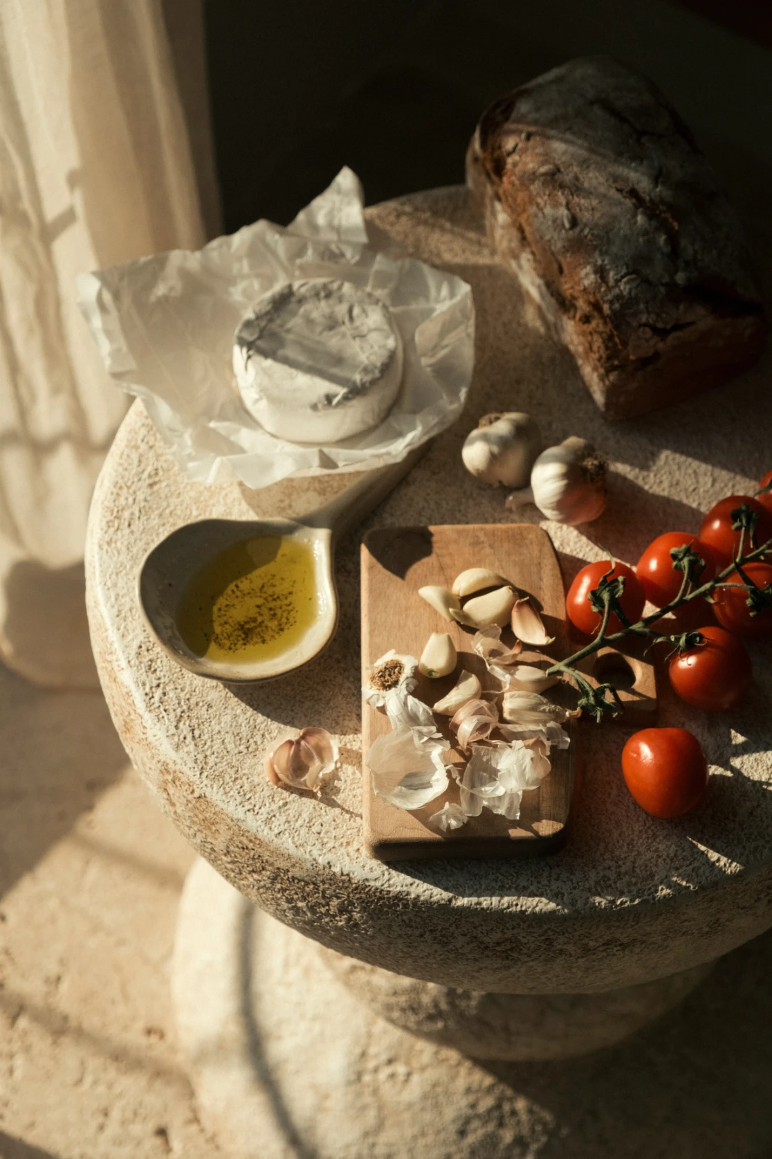 Garlic cloves on a wooden board with vine tomatoes, a wrapped soft cheese, and olive oil on a stone table.