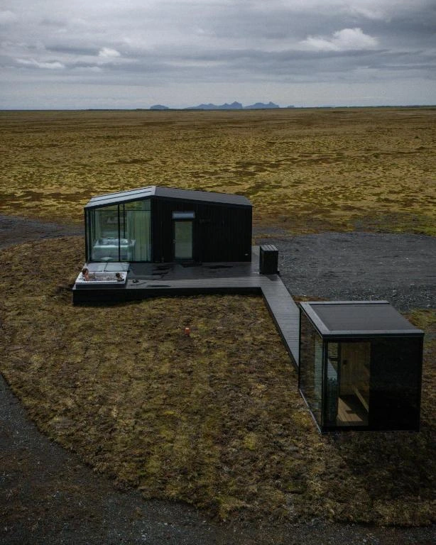 A dark timber cabin with an outdoor hot tub and glass pod at Harmony Retreat Lodge, set on open Icelandic moorland.