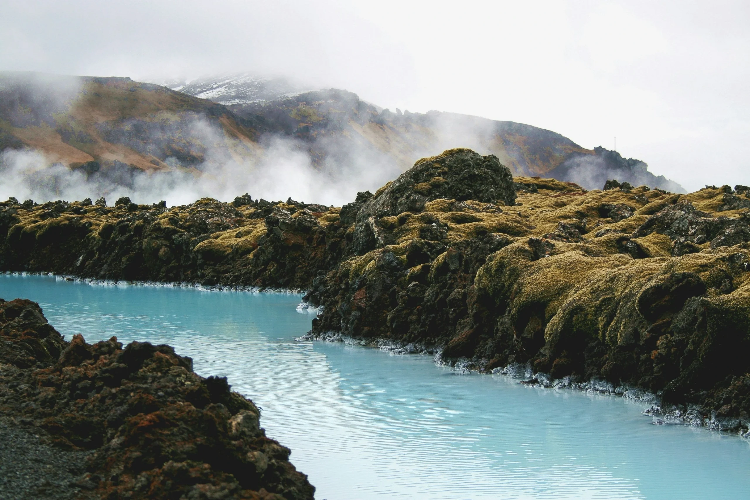 Milky blue geothermal water winds through moss-covered lava rock in Iceland, with steam drifting over the hills.
