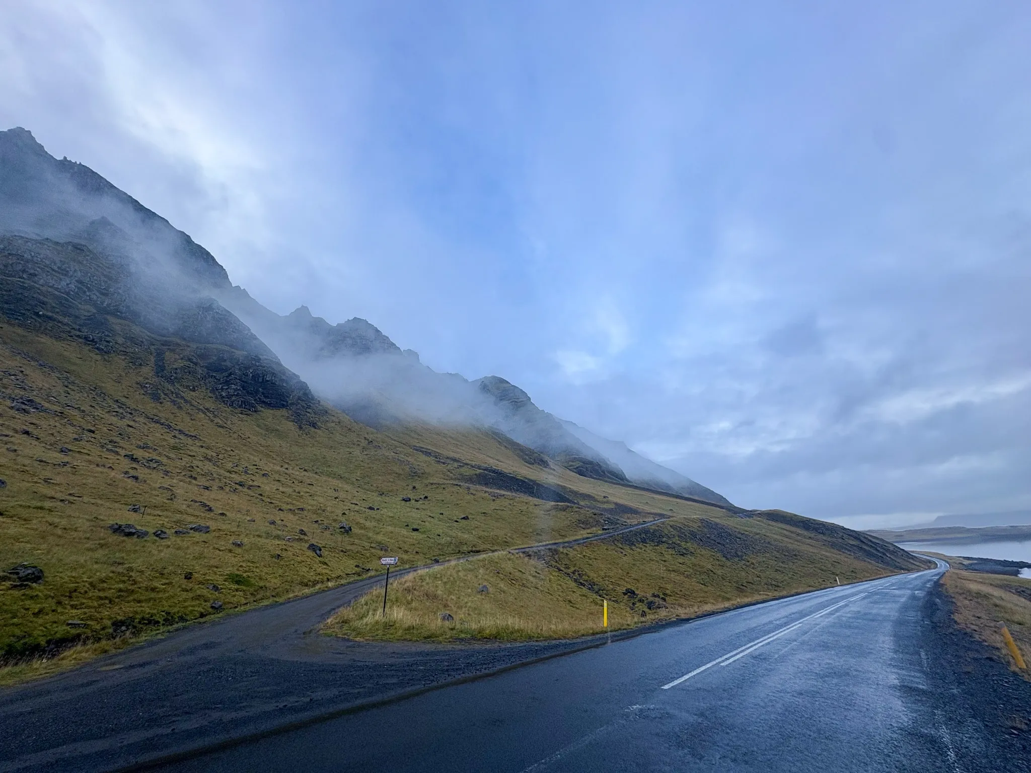 A rain-slicked road winds beneath mist-wrapped mountains toward a fjord in Iceland.