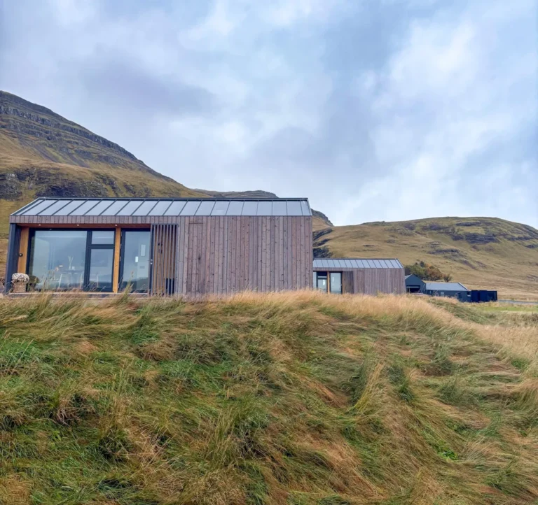 EYRI Farm cabins with wide windows sit beneath grassy mountain slopes in the Icelandic countryside.