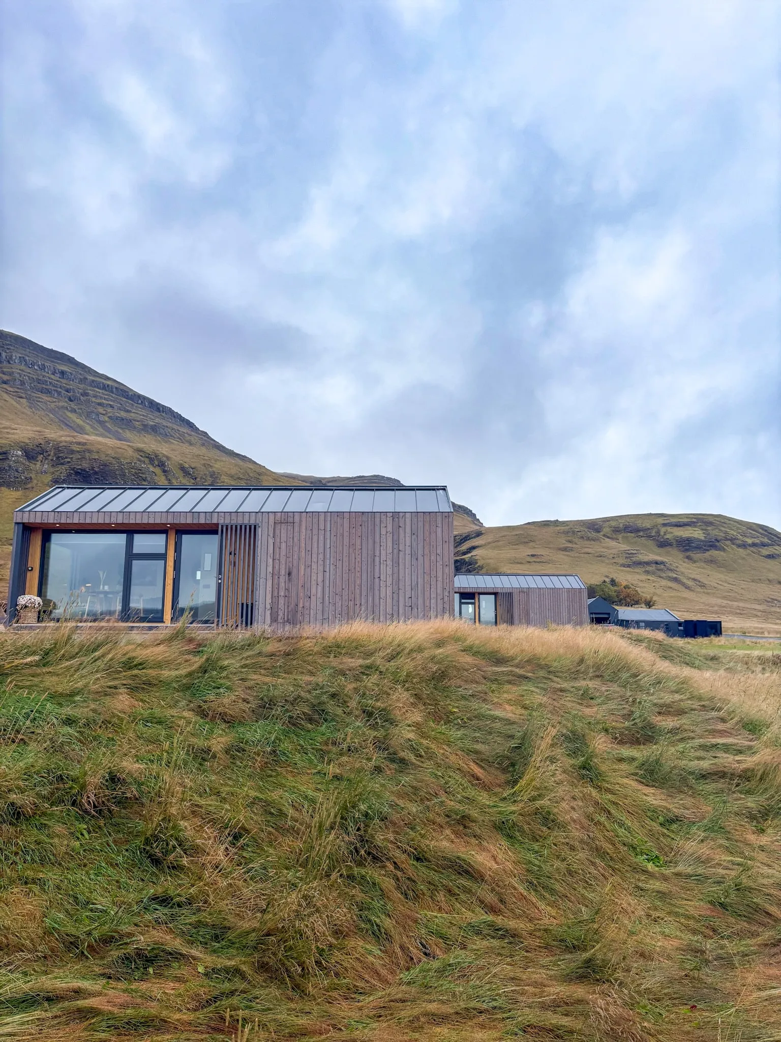 Timber-clad cabins with wide windows sit beneath grassy mountain slopes in the Icelandic countryside.