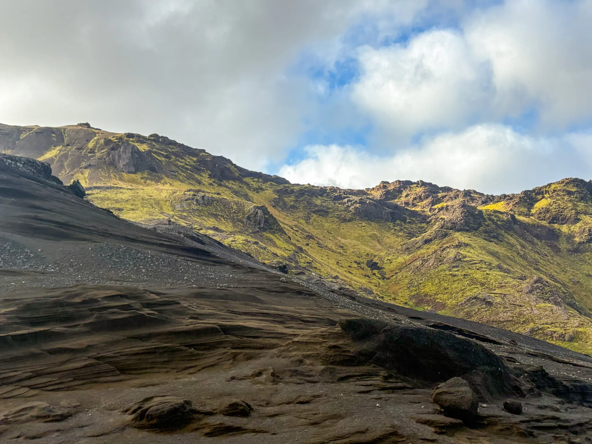 Wind-carved volcanic rock and dark ash slopes lead toward moss-green mountains under drifting cloud in Iceland.