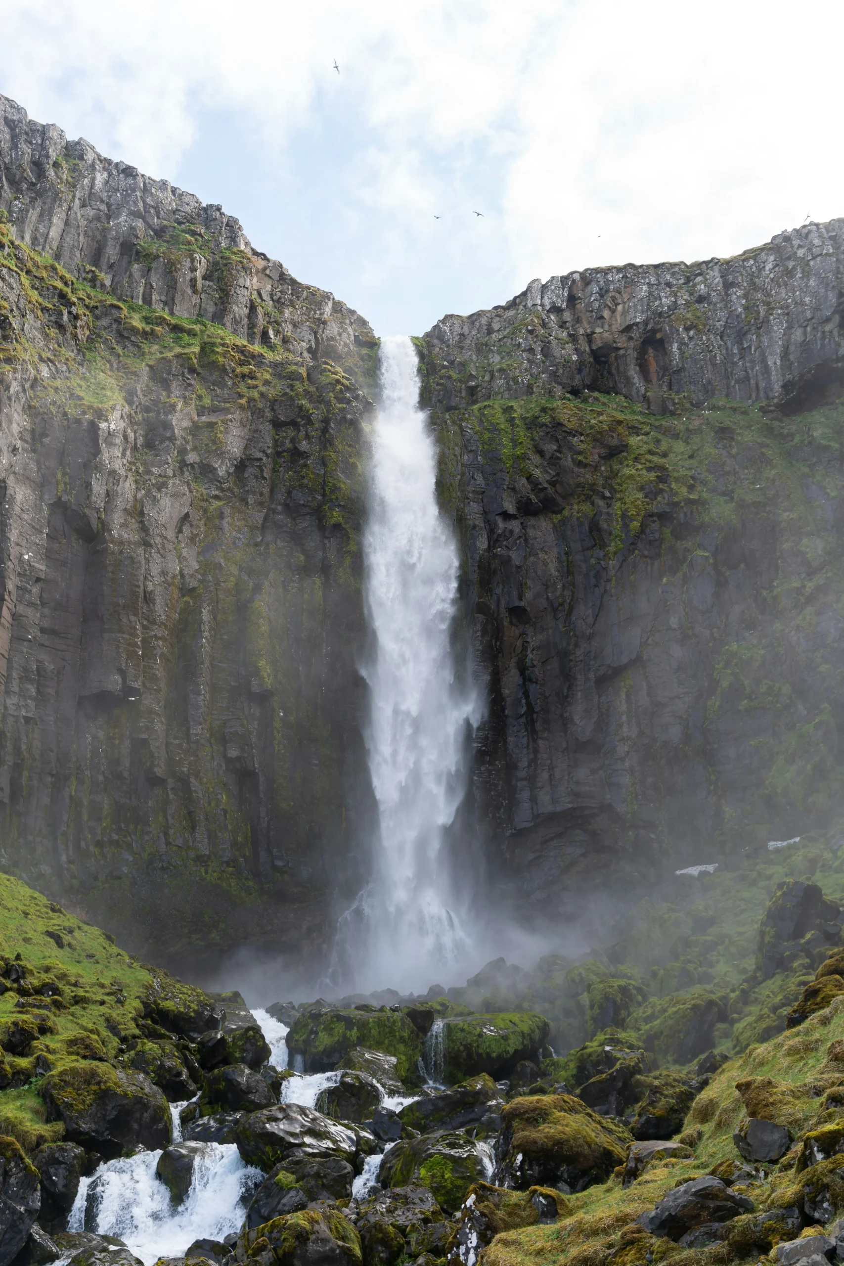 A tall waterfall plunges between moss-covered basalt cliffs into a rocky gorge in Iceland.
