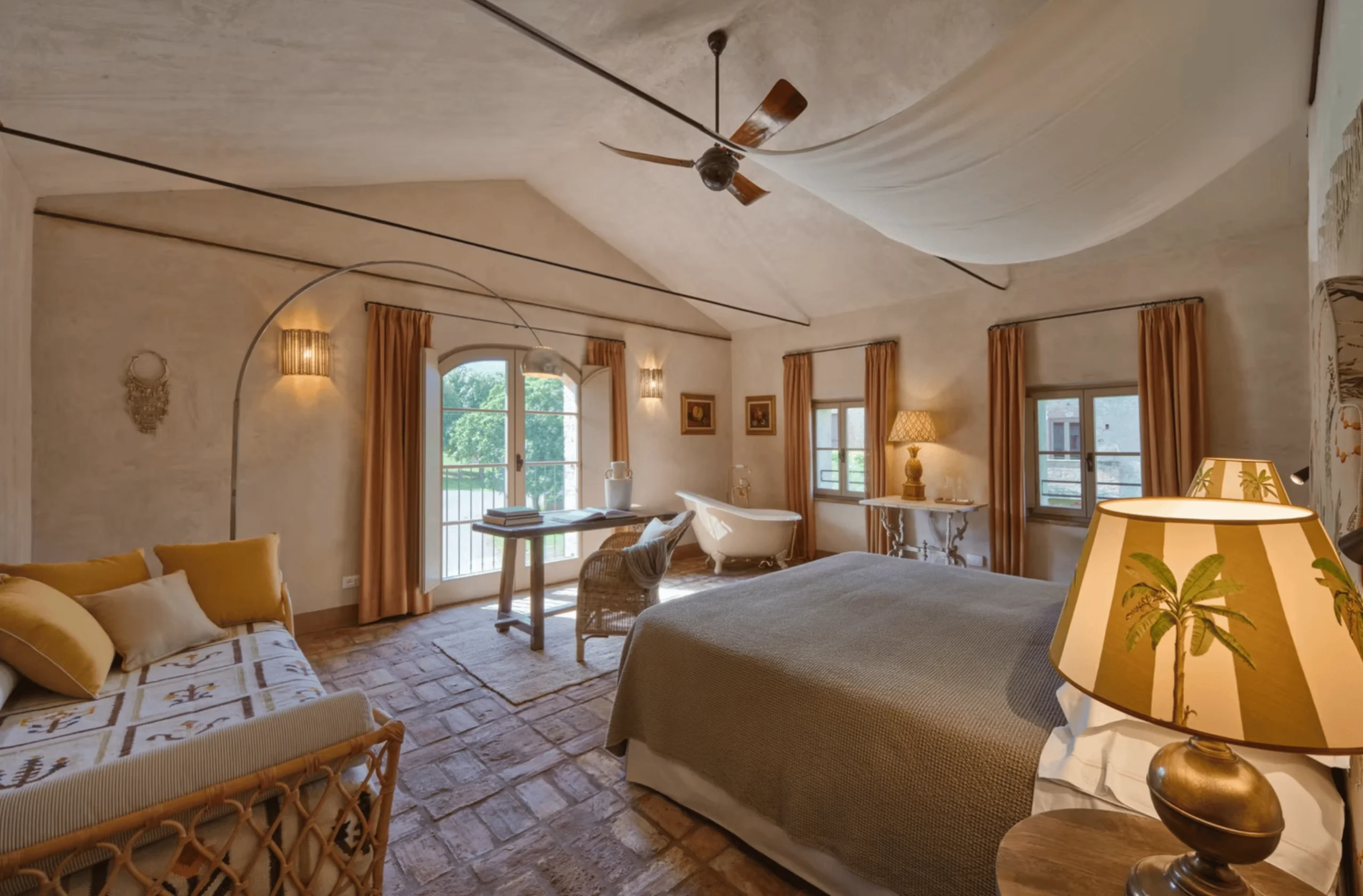 A vaulted bedroom at La Pescaia with a linen canopy, clawfoot tub, rattan daybed, and an arched window to the garden.