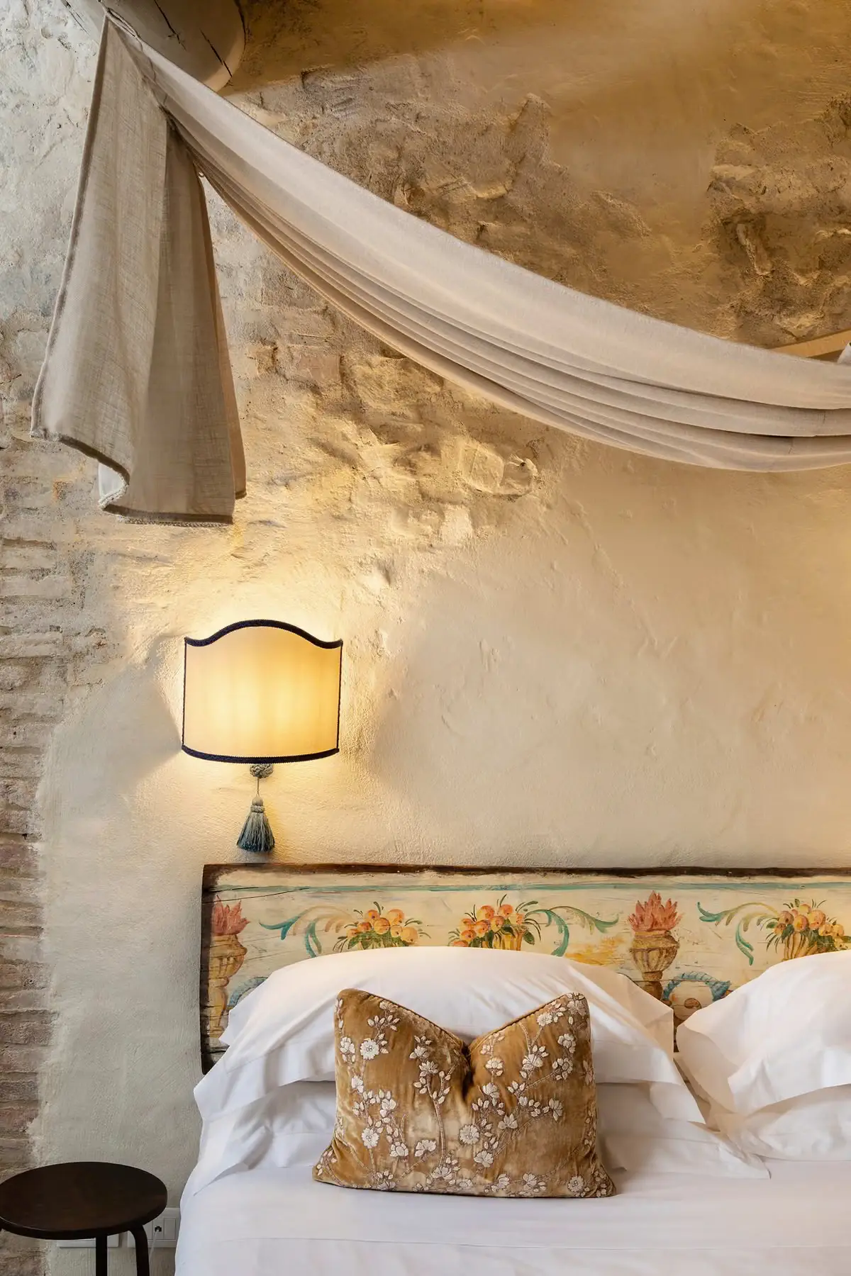 A hand-painted floral headboard beneath a draped linen canopy at Lupaia, with a scalloped sconce on a stone wall.