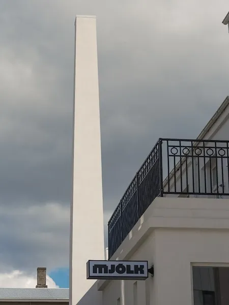 The white facade and slender chimney of Mjólk hotel, with its signage below a wrought-iron balcony.