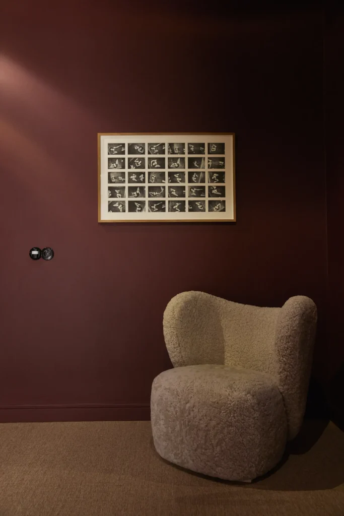 A shearling armchair below a framed photographic contact sheet against a deep burgundy wall at Mjólk hotel.