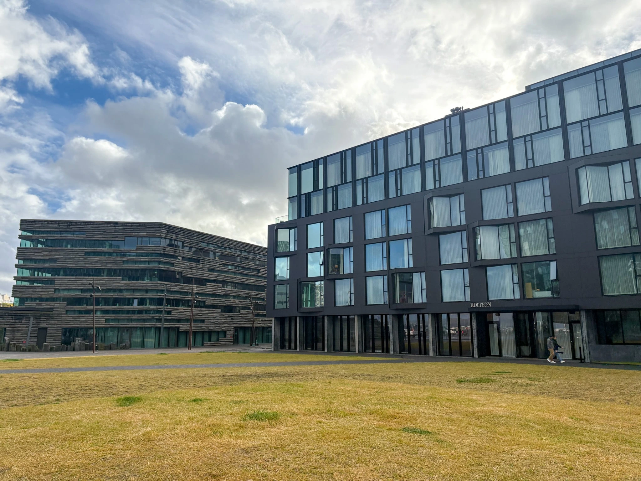 The dark glass facade of The Reykjavik EDITION hotel, set beside a broad lawn beneath a cloud-filled sky.