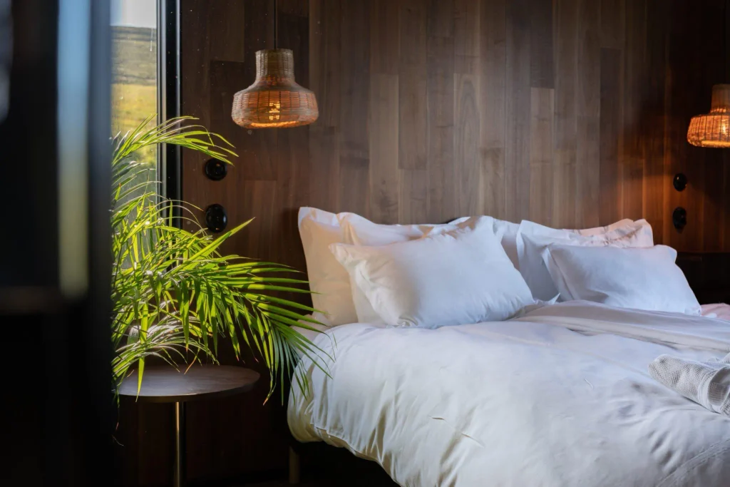 White bedding beneath rattan pendants and walnut paneling, with a palm leaning in at Svartaborg Luxury Villa.
