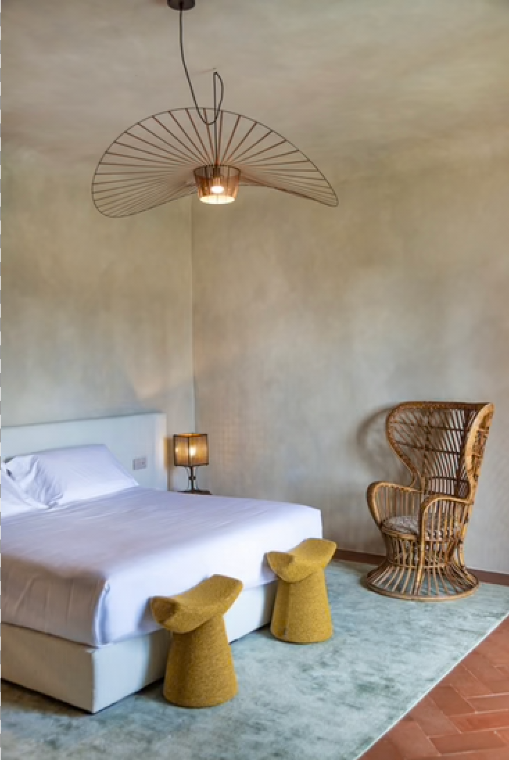 A plaster-walled bedroom at Wine Relais Ruffino with a wire pendant lamp, wicker peacock chair, and mustard stools.