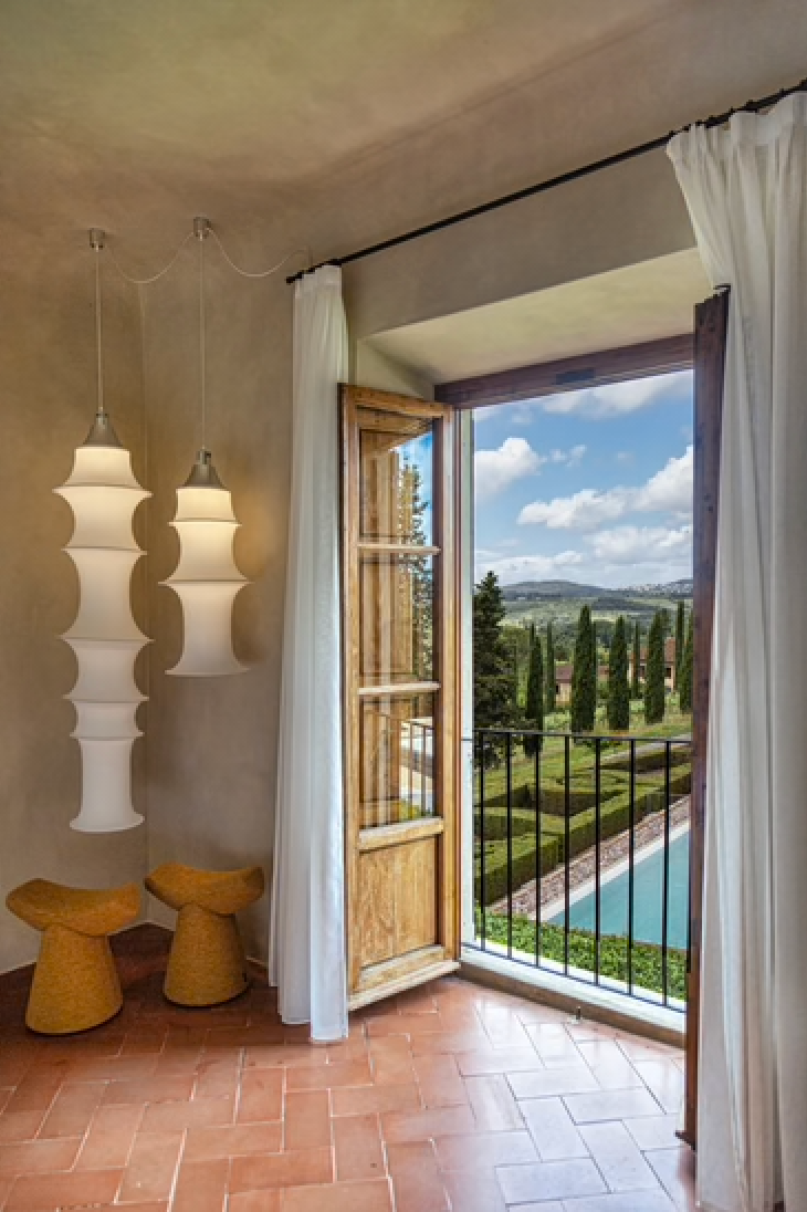Open shutters at Wine Relais Ruffino frame a view of box-hedge gardens, a pool, and cypresses beside paper lanterns.